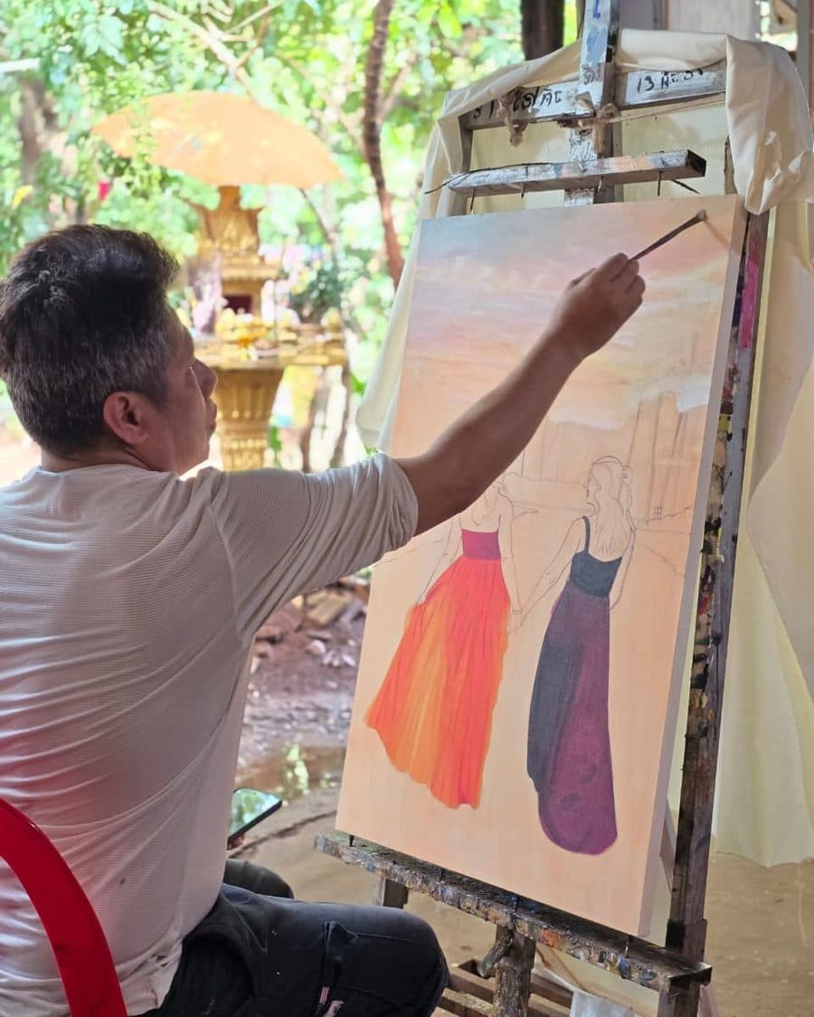 A Velea artist at the easel, brush raised to the canvas, working from a customer photograph in oil.